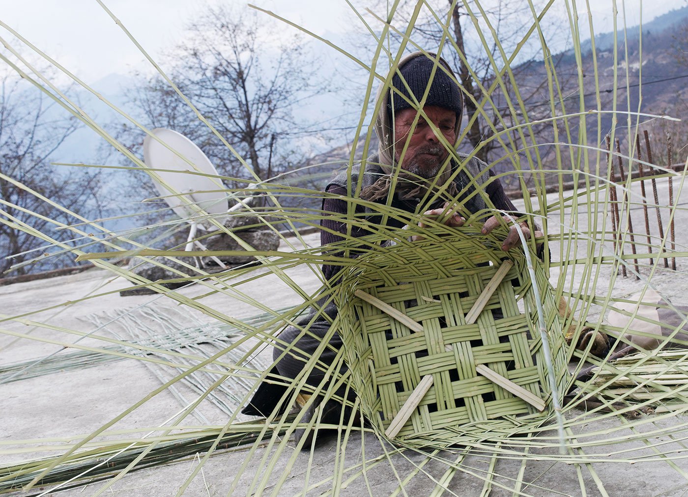 A man sitting on a terrace of a house and weaving bamboo strips into a cylindrical shape