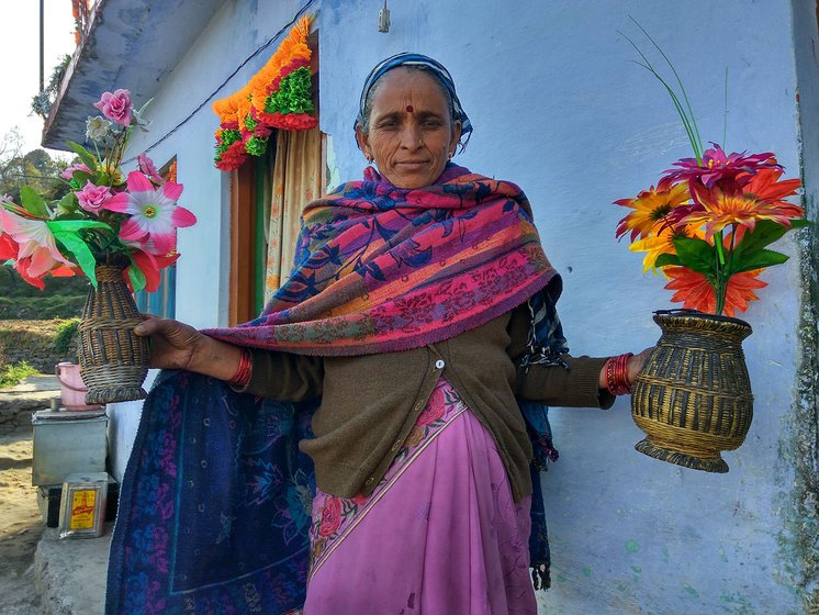 A woman standing outside her house holding two woven bamboo vases filled with plastic flowers